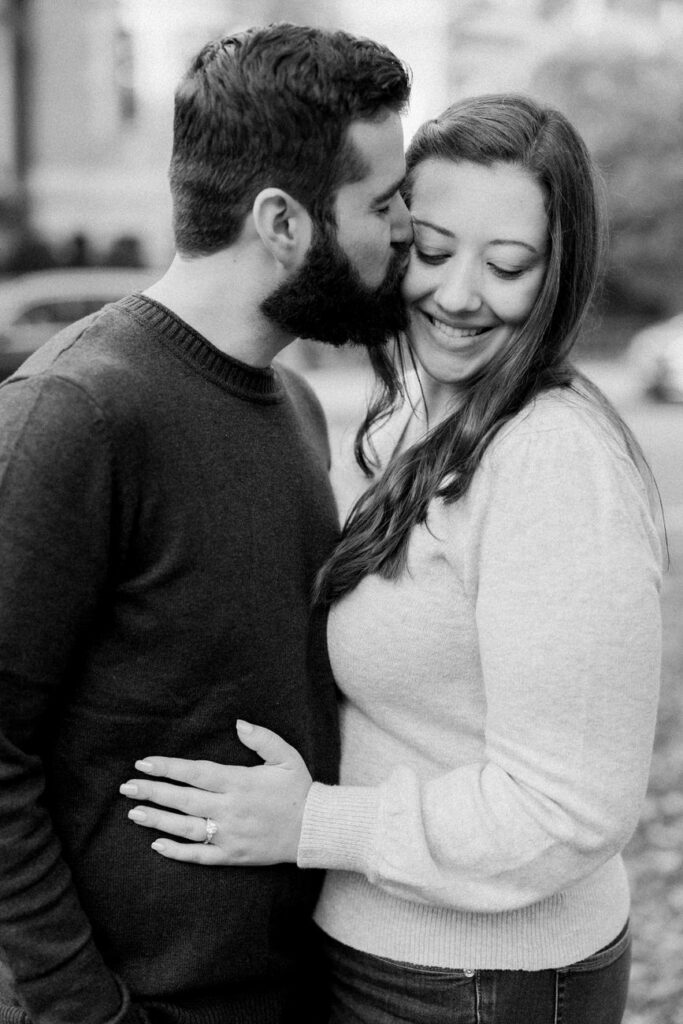 A tender moment in monochrome where a bearded man kisses his partner's forehead, both smiling gently, in an urban setting.