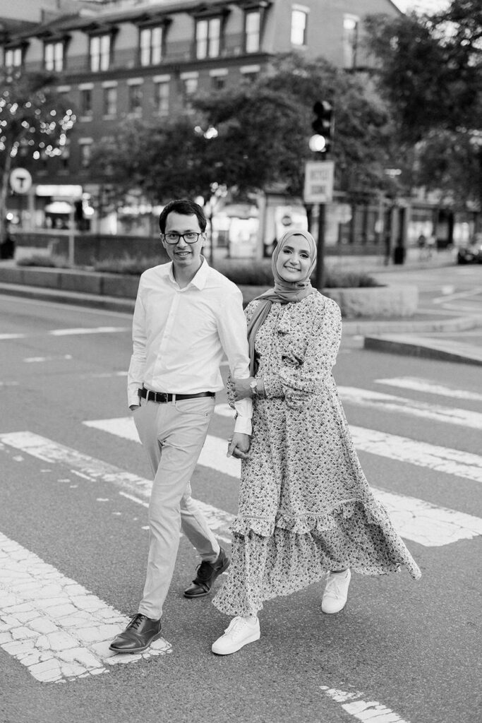 Black and white photo of a couple crossing the street, the woman in a flowing patterned dress and headscarf, the man in a casual white shirt and dark pants.