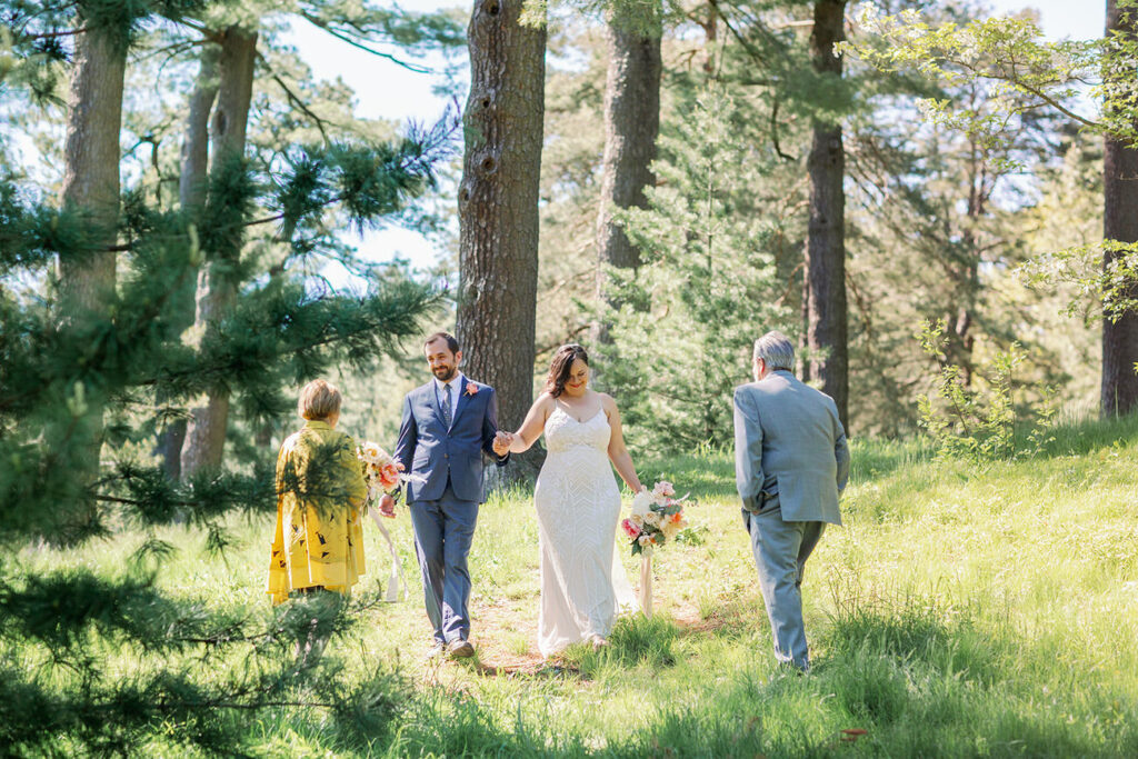 A cheerful bride and groom walk hand in hand through a lush pine forest, with an officiant and another guest trailing behind, capturing the beauty of a serene outdoor wedding.