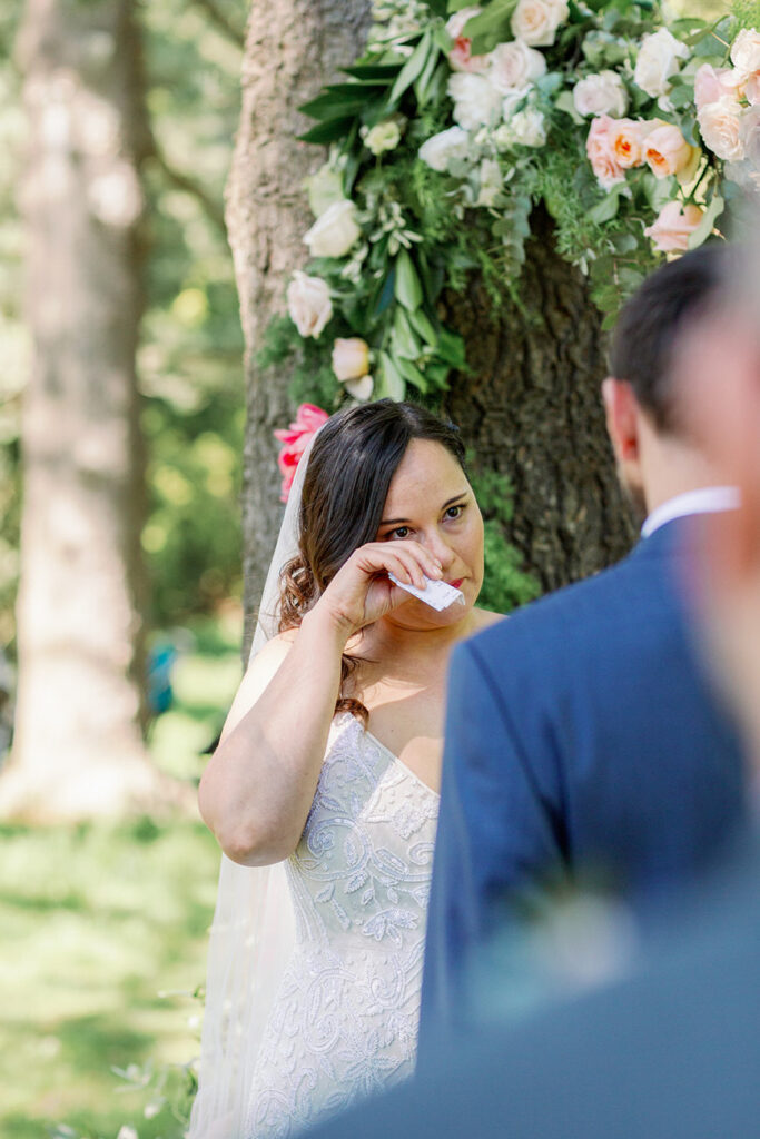The bride, clad in an elegant white lace gown, holds a tissue to her eyes in a moment of emotion, with a beautifully detailed floral arrangement adorning the tree beside her.