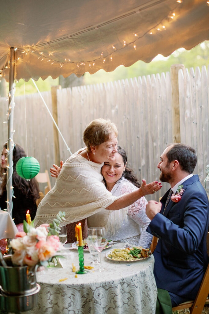A heartwarming evening scene under a canopy of fairy lights where a bride embraces a guest, possibly a family member, at a festively decorated wedding reception table.
