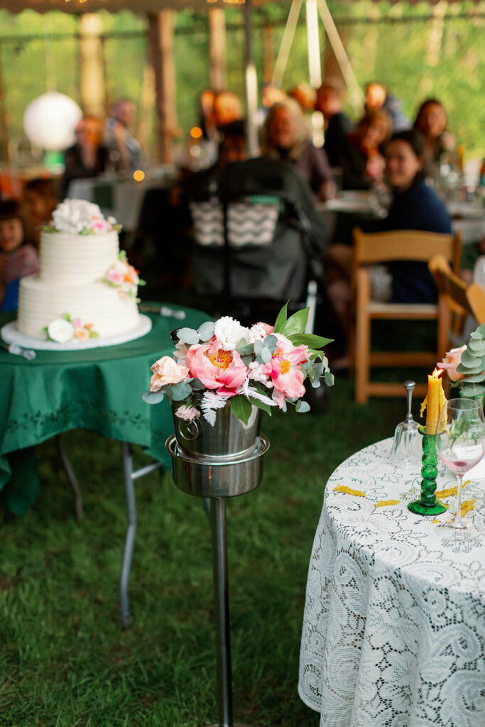 A wedding celebration's outdoor reception area illuminated by warm light, featuring a tiered white cake adorned with flowers, set next to a table with elegant green candles and floral decorations