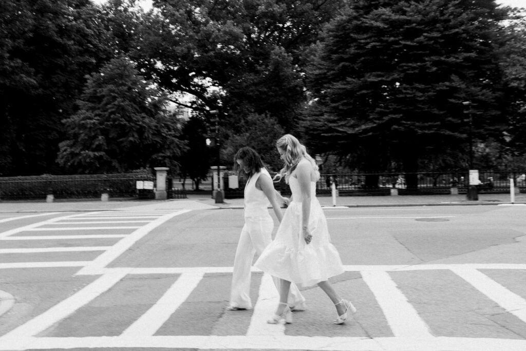 A timeless black-and-white image capturing two women in wedding dresses, joyfully crossing a city street.