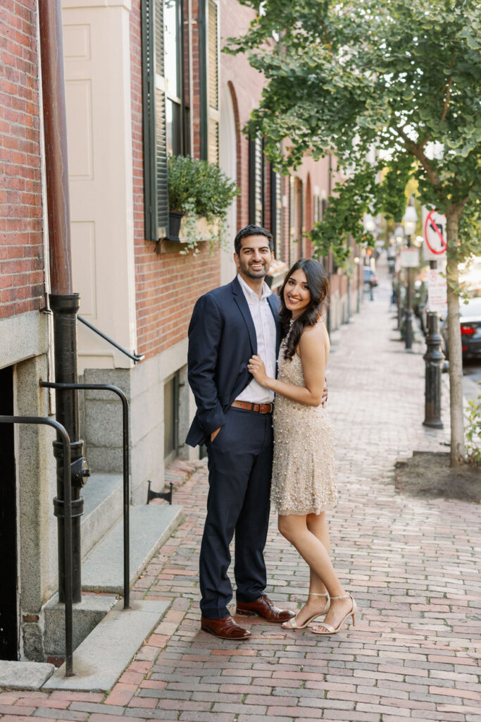 A radiant couple stands arm in arm on a brick-lined sidewalk in Boston's Beacon Hill, with historic brownstone buildings and leafy trees framing the charming scene.