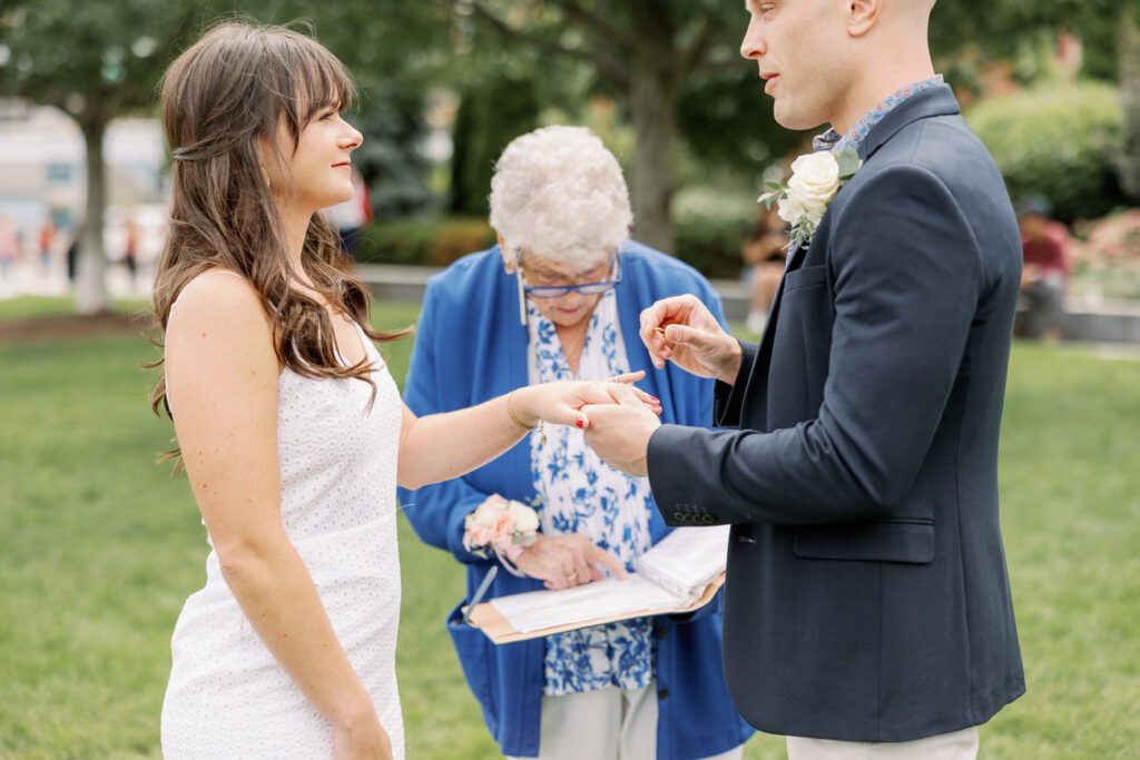A woman in a white dress and a man in a suit exchanging wedding rings, with an officiant in blue assisting them, during an intimate outdoor ceremony.