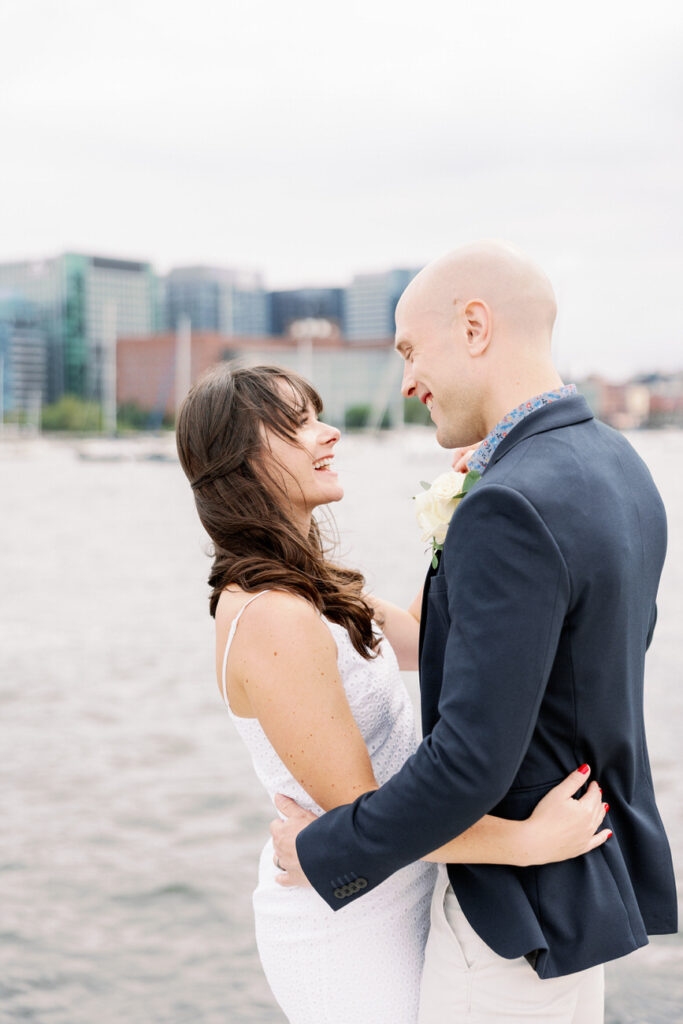 A newlywed couple embraces near the waterfront, with the Boston skyline behind them, capturing a moment of joy and love.