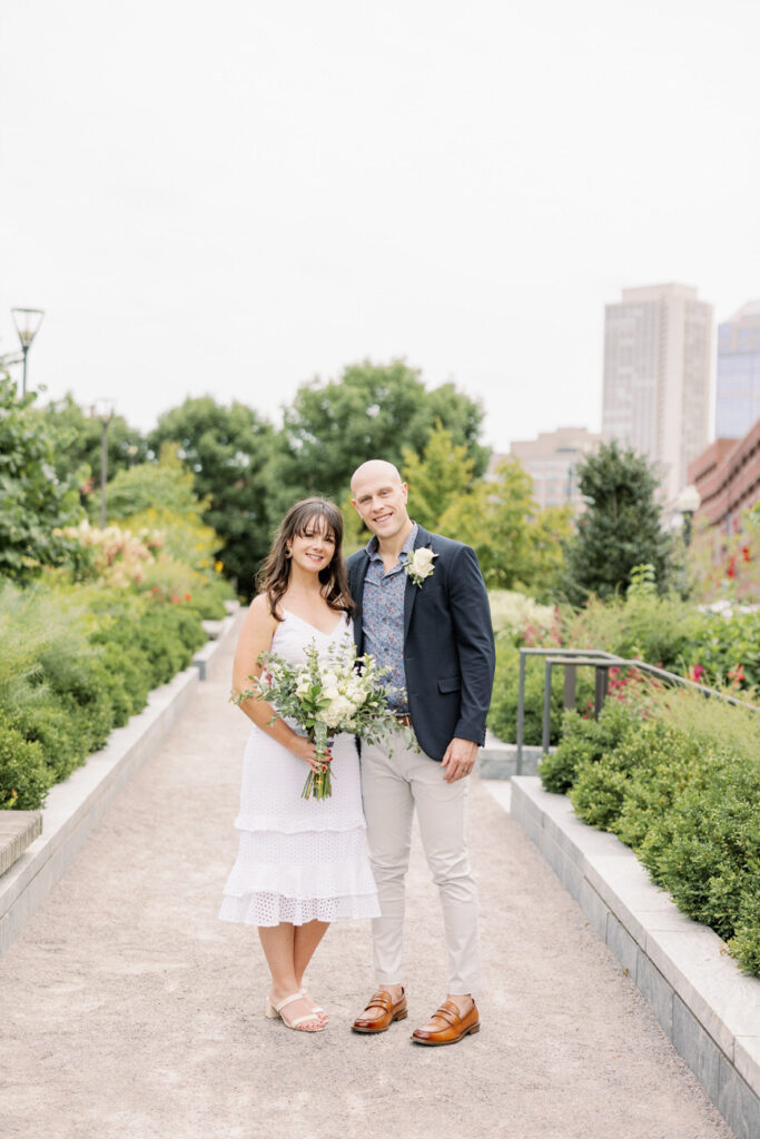 A bride and groom walk hand-in-hand along the Boston Public Garden bridge, surrounded by lush greenery and the cityscape.