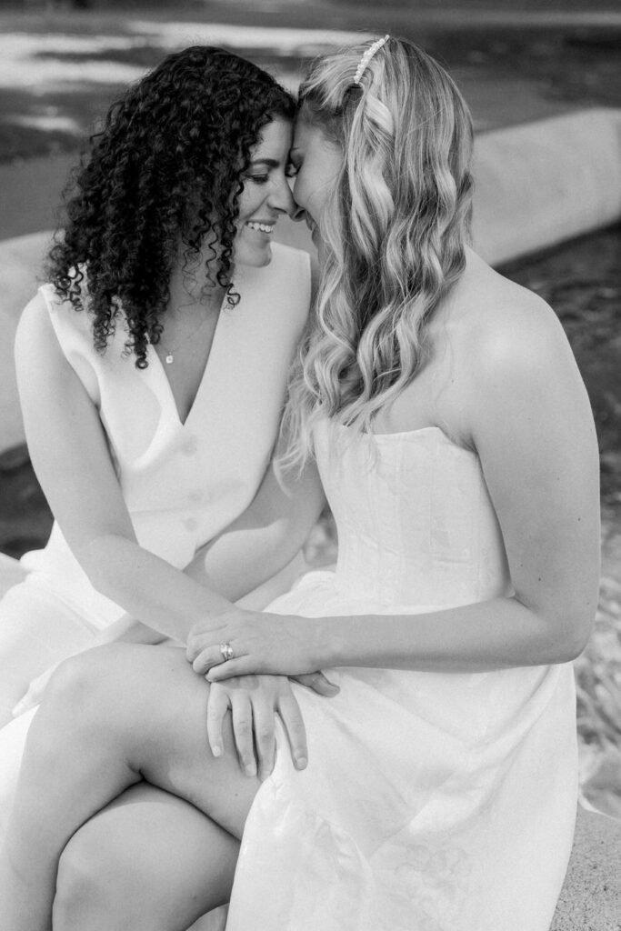 A black and white photo capturing an intimate moment between two women in wedding dresses, seated and sharing a quiet moment together.