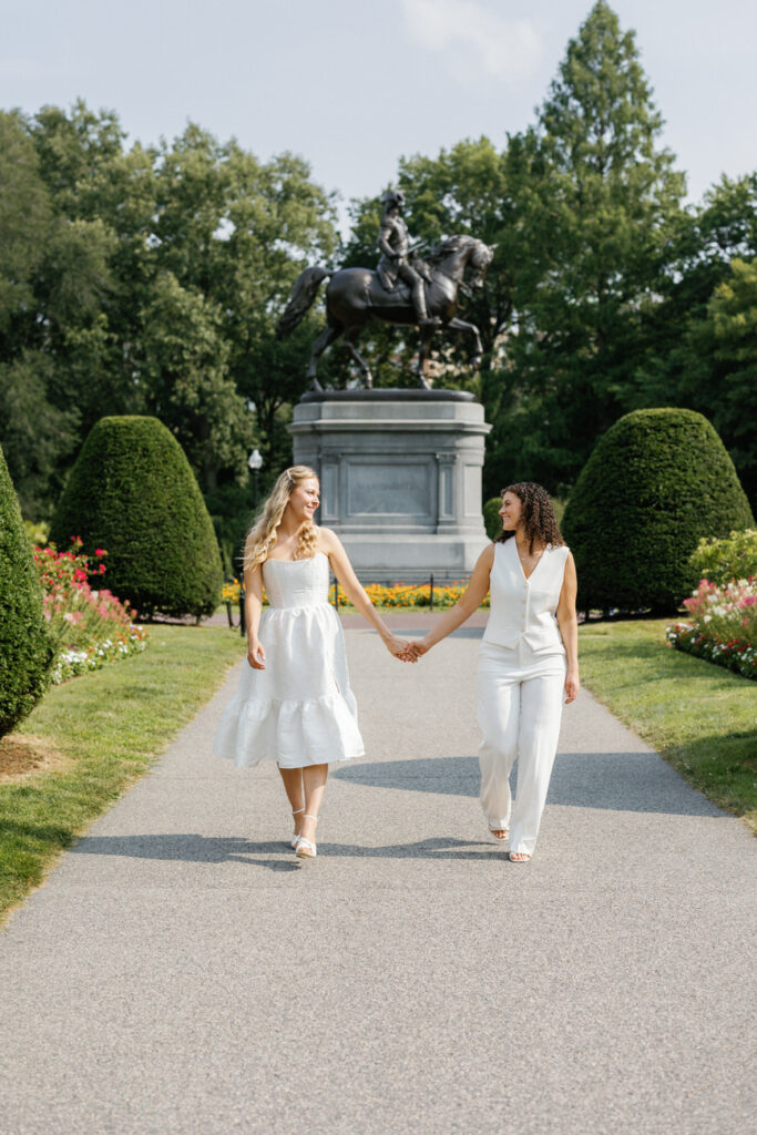 Two brides walk towards the camera, holding hands, with a statue and vibrant garden in the background.