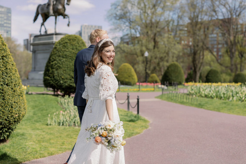 A bride and groom walk along a path in the Boston Public Garden, the bride holding a bouquet, with the statue in the background.
