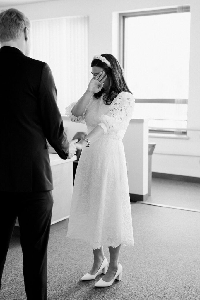 A bride in a vintage lace tea-length dress with a headband cries joyfully, holding the hand of a person in a dark suit, in a candid black and white photo