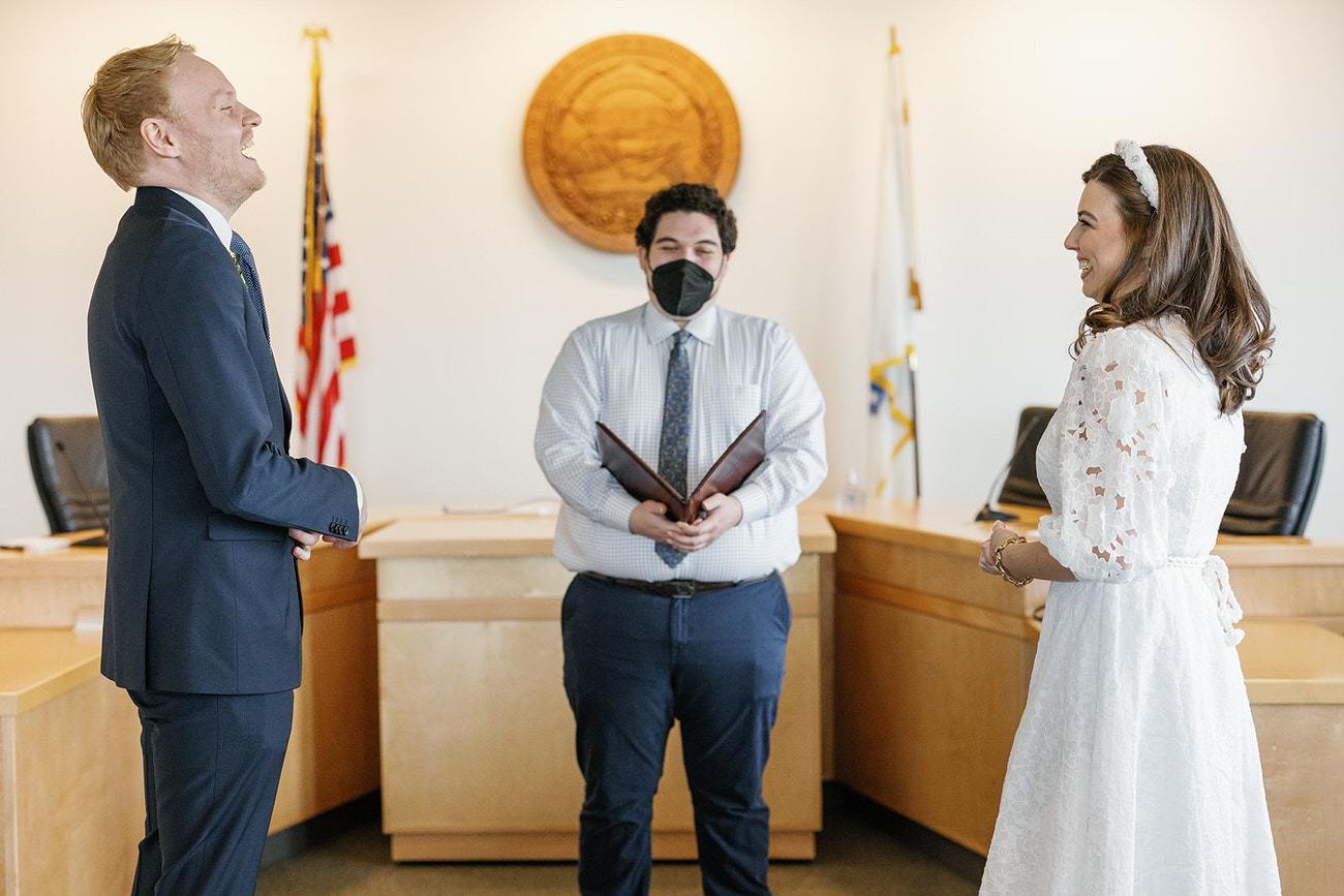 A couple laughs heartily in a courtroom setting, the bride in her lace wedding dress and the groom in a navy suit, with an officiant holding a brown folder in the background