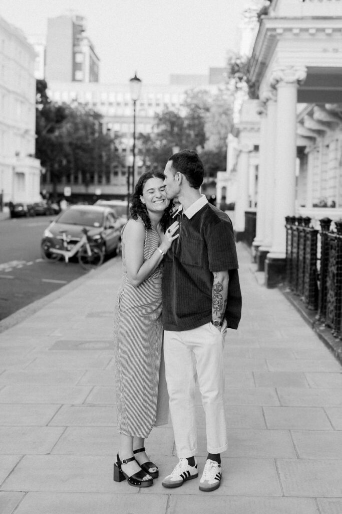 Monochrome image of a couple standing close and smiling on a city sidewalk, with a classic London street and architecture in the background.