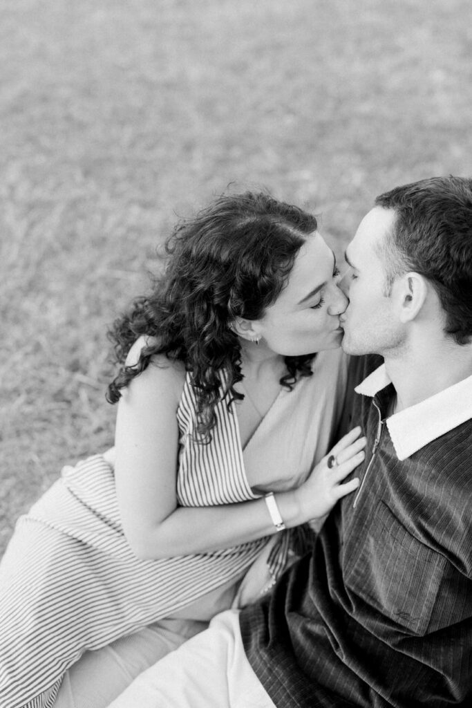 A black and white photo capturing an intimate moment of a couple kissing while sitting on a grassy field, with the man's arm around the woman.