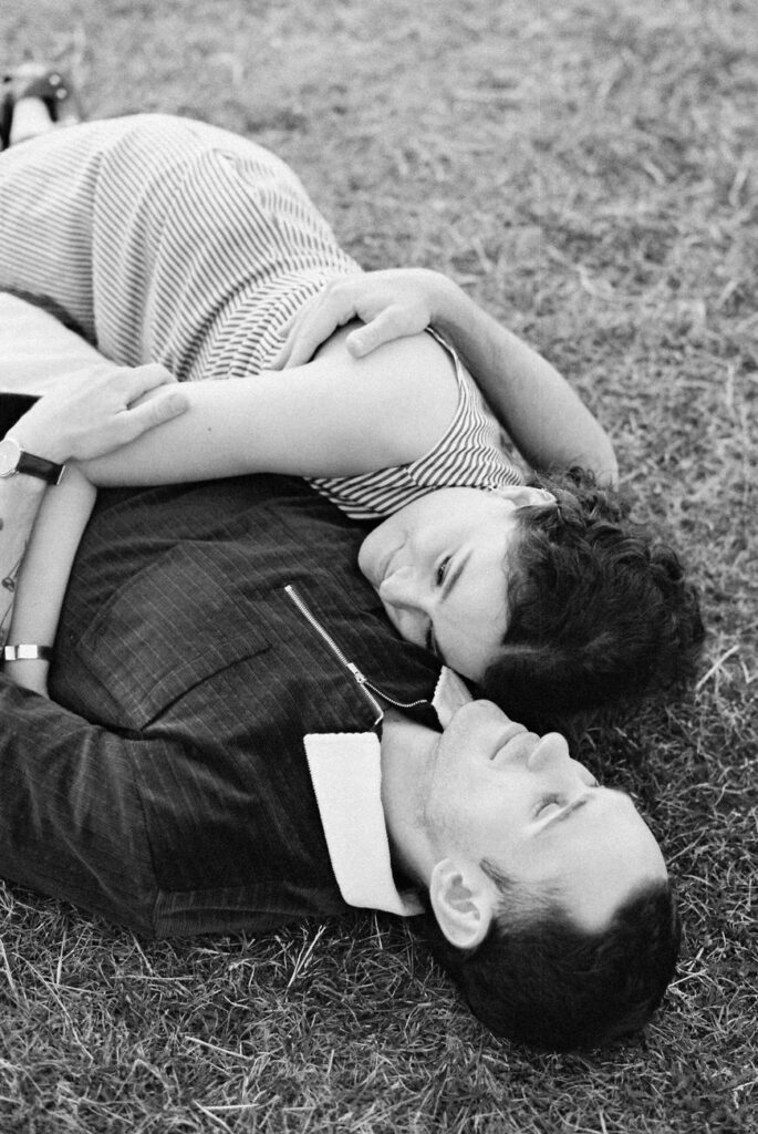 An affectionate black and white image of a couple lying on the grass, embracing each other, with a relaxed and content expression on their faces.