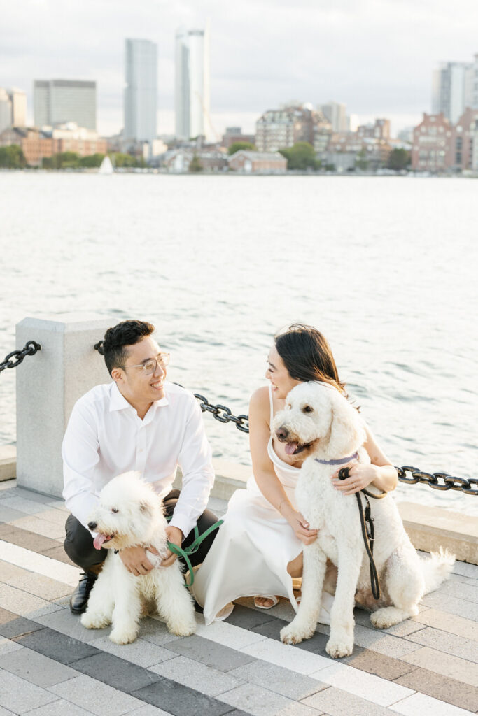 A couple seated by the Boston waterfront, smiling and looking at each other, with two fluffy white dogs by their side and city buildings blurred in the background.