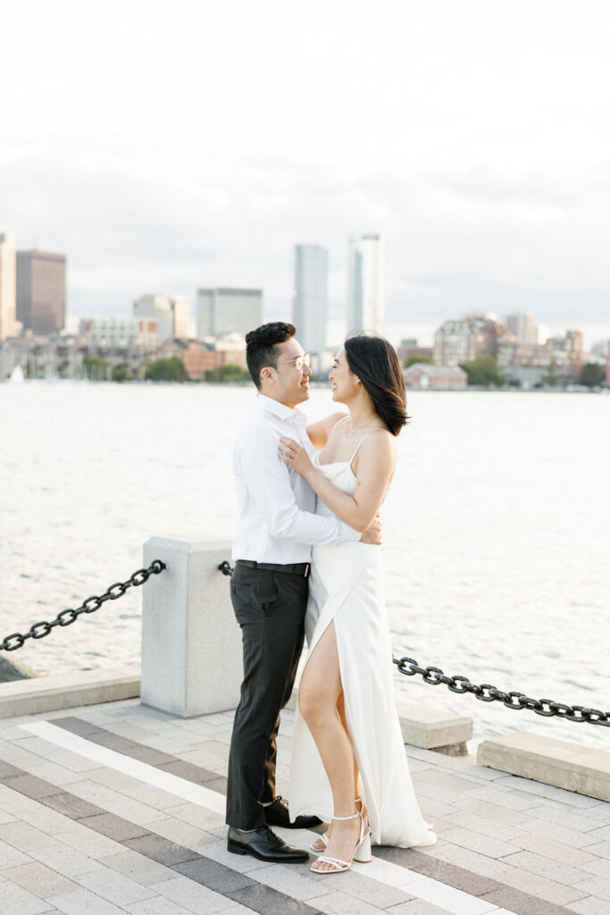 A couple stands close together, embracing on the waterfront, with the Boston skyline in the background, creating a romantic city scene.