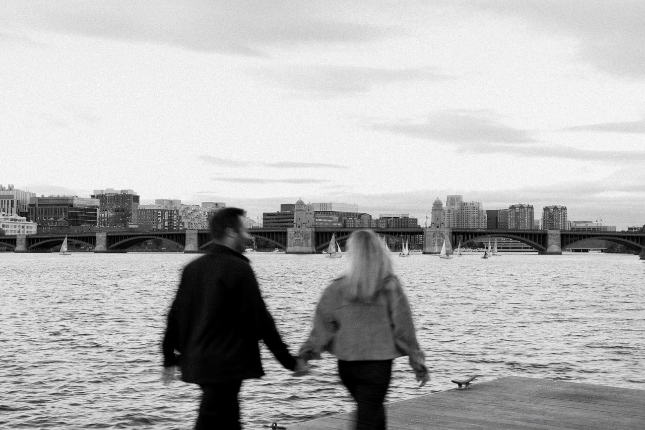 Monochrome photo showing a couple walking hand in hand along a waterfront, with sailboats on the water and a cityscape in the distance.