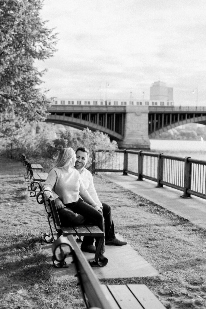 A black and white image of a couple seated on a park bench under a tree, smiling and looking at each other with a bridge in the background