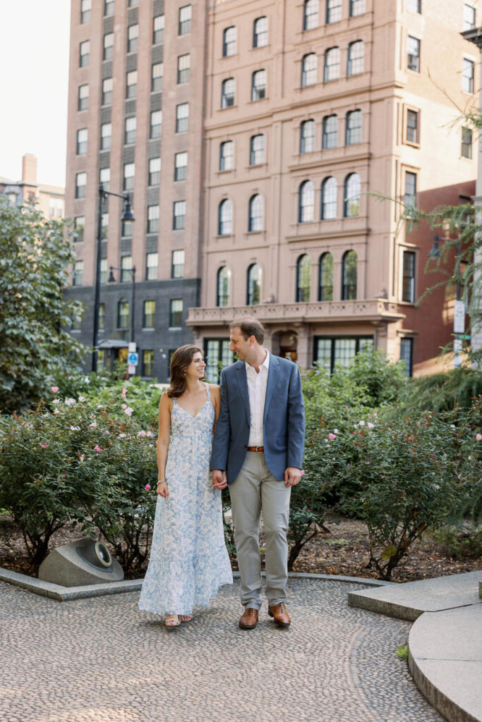 A couple strolling hand in hand, looking at each other lovingly, in a serene park with lush greenery and urban buildings in the background.