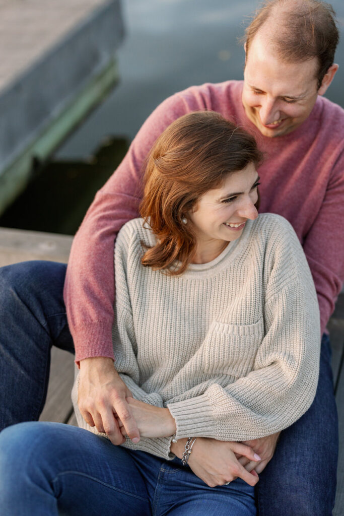 An intimate moment with a couple seated on a dock, the woman in a cozy sweater leans on the man's shoulder, surrounded by tranquil waters.