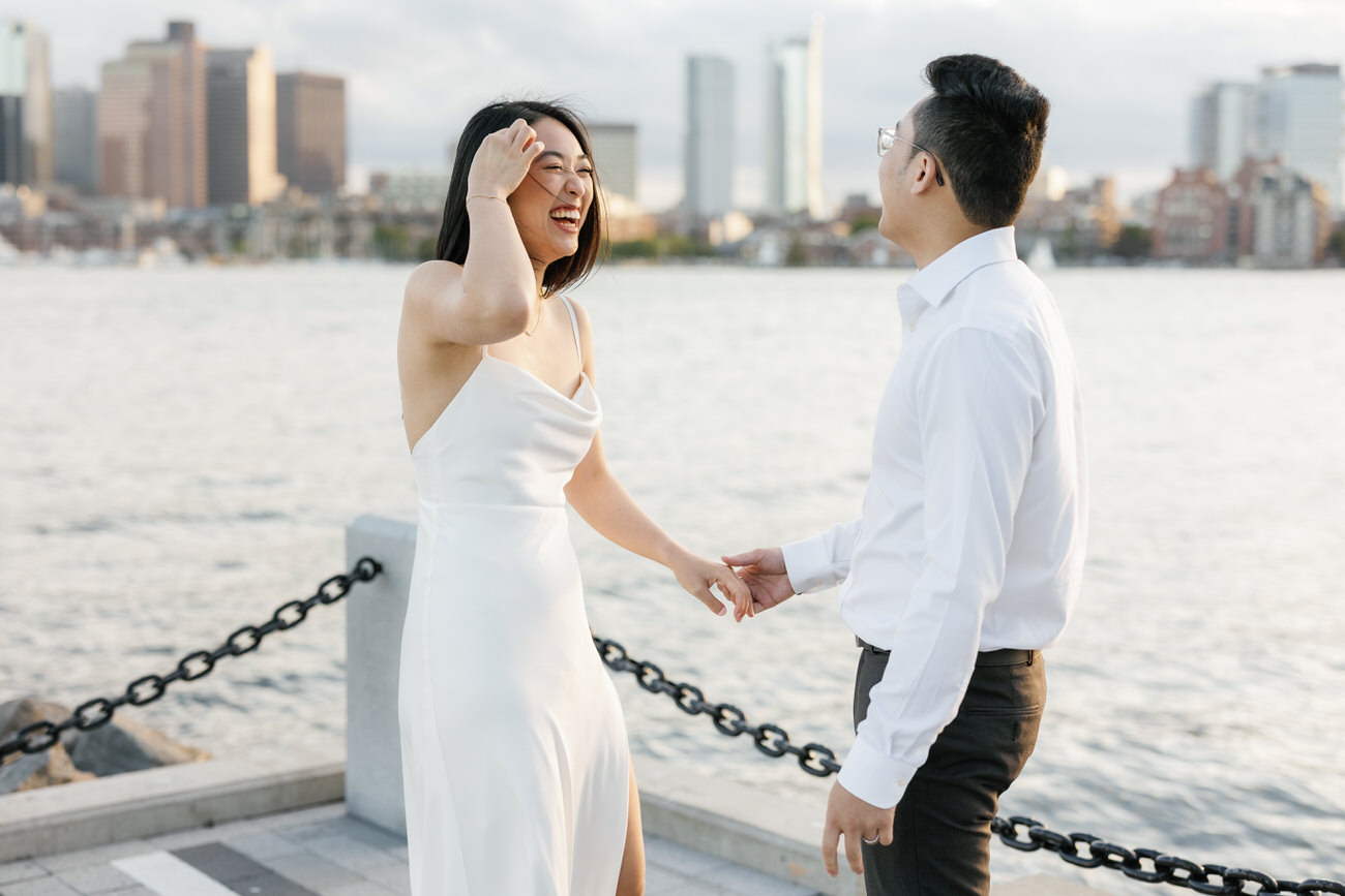 A joyful woman in a white dress laughing with her partner by the waterfront, cityscape glowing in the soft light of dusk.