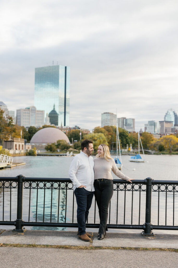 A romantic couple standing close, with a city riverfront and autumnal trees in the soft glow of the setting sun as their backdrop.