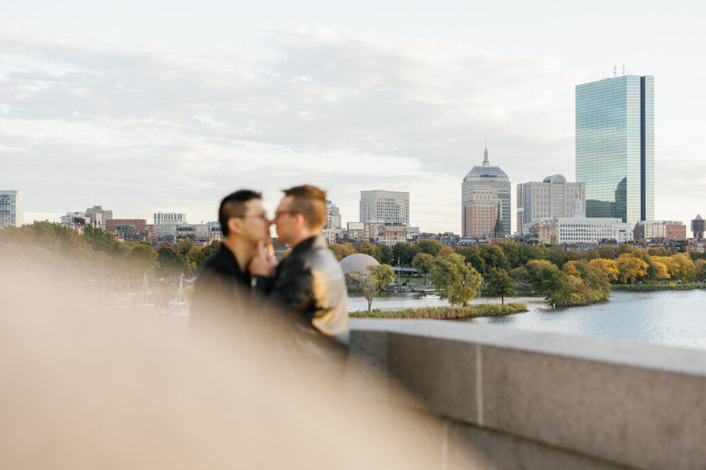 An affectionate moment with a couple kissing, partially obscured by foliage, city architecture forming a picturesque background.