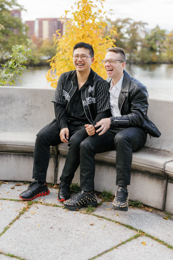 Two men in stylish black jackets share a laugh while sitting on a park bench, enjoying a vibrant autumn day.