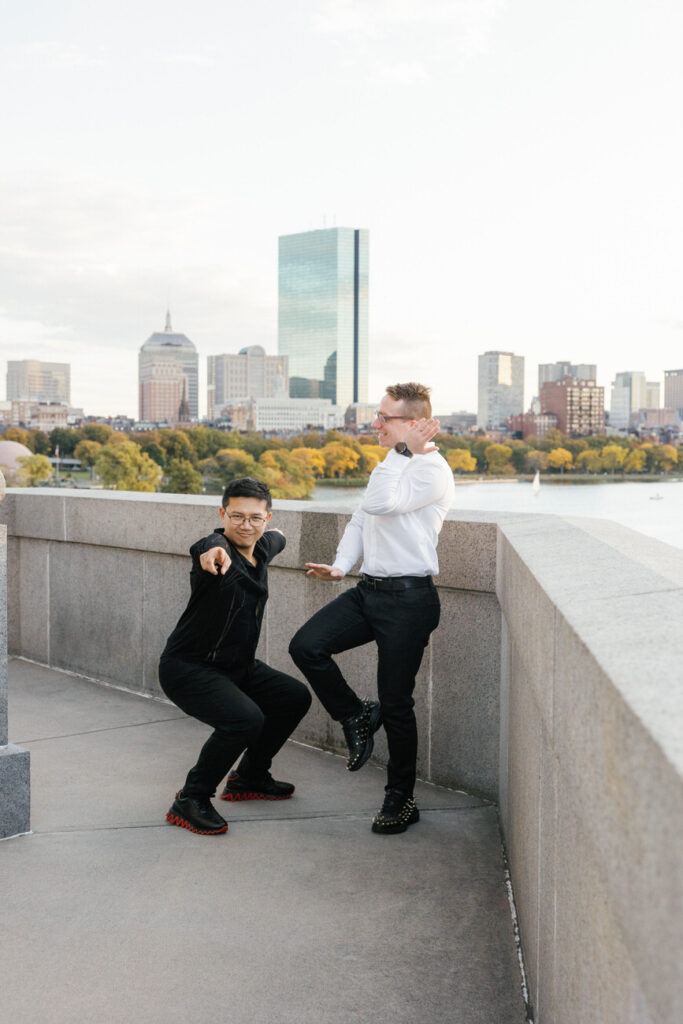 Two lovers sharing a playful moment on a city overlook, one squatting and pointing forward, the other standing with hand on hip, with a soft-focused city skyline in the background.