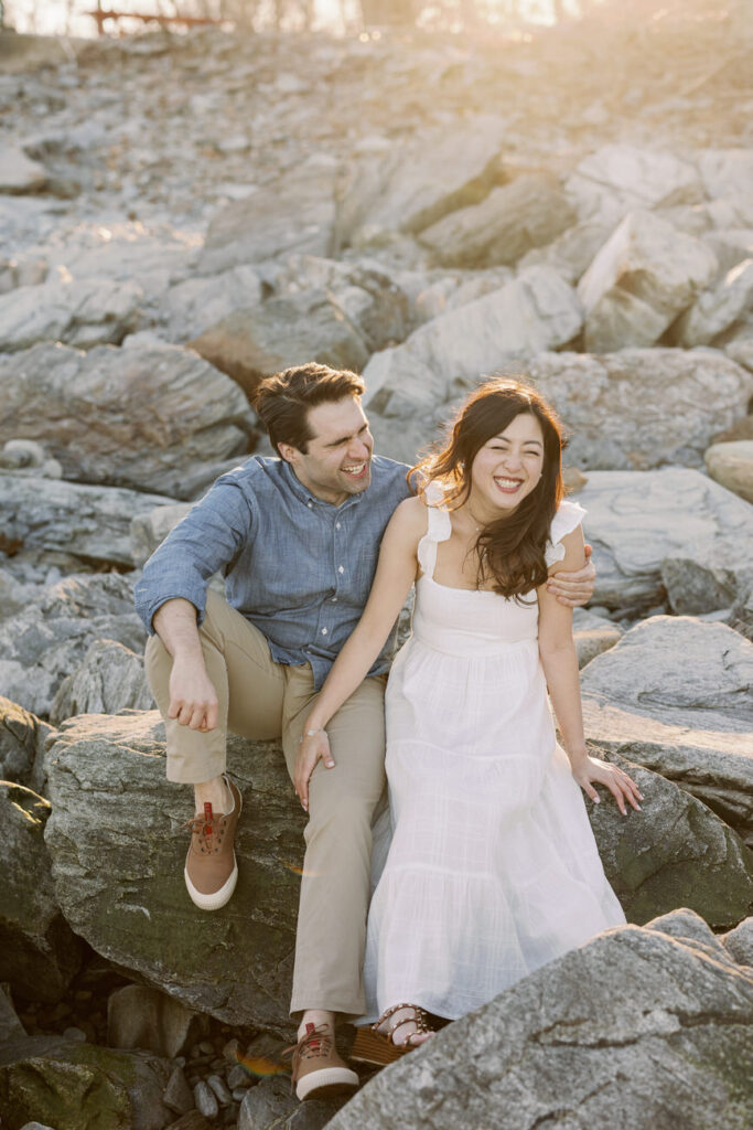 A couple seated on rugged coastal rocks, sharing laughter in the warm glow of the setting sun, with the woman's dress flowing in the gentle sea breeze.