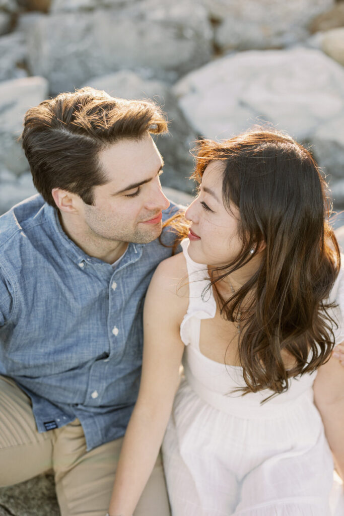An intimate close-up of a couple on the rocks, the man in a denim shirt leaning in towards the woman in a white dress, with a soft-focus on their smiling faces.
