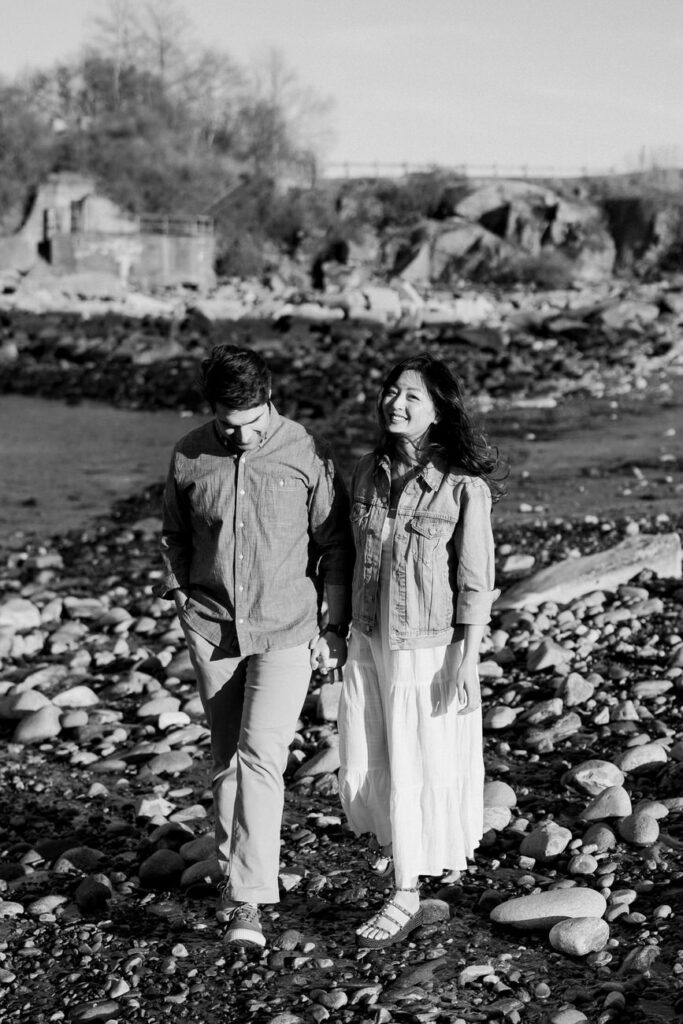A couple walking on a pebble-covered shore, the man in a denim shirt and the woman in a flowing dress, both looking off into the distance with smiles.