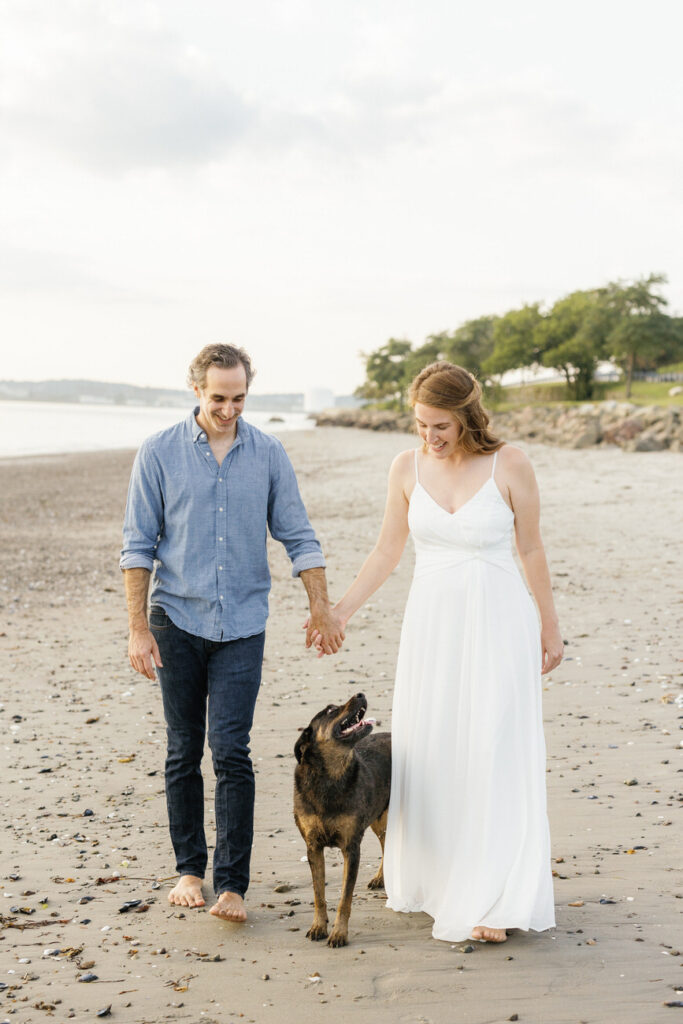 A couple walking on the beach, the man in a casual blue shirt and the woman in a white dress, with a happy dog leading the way and looking back at them with a playful stance.