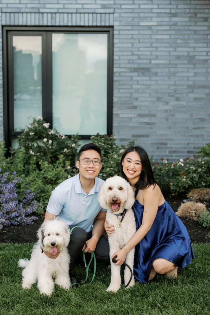 A smiling couple seated on the grass with their two adorable dogs; one dog is a fluffy white and the other a cheerful goldendoodle, with a modern home's gray brick facade and windows in the background.