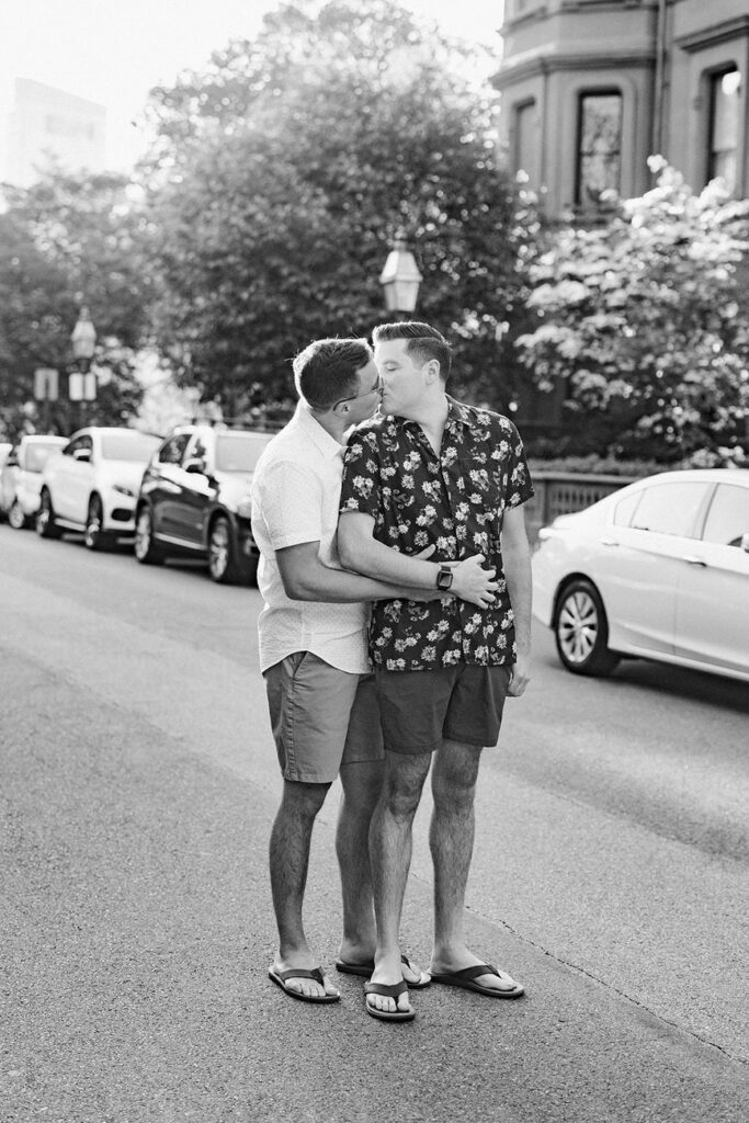 A grayscale image capturing a moment between two men embracing and touching foreheads on a sunny city street.