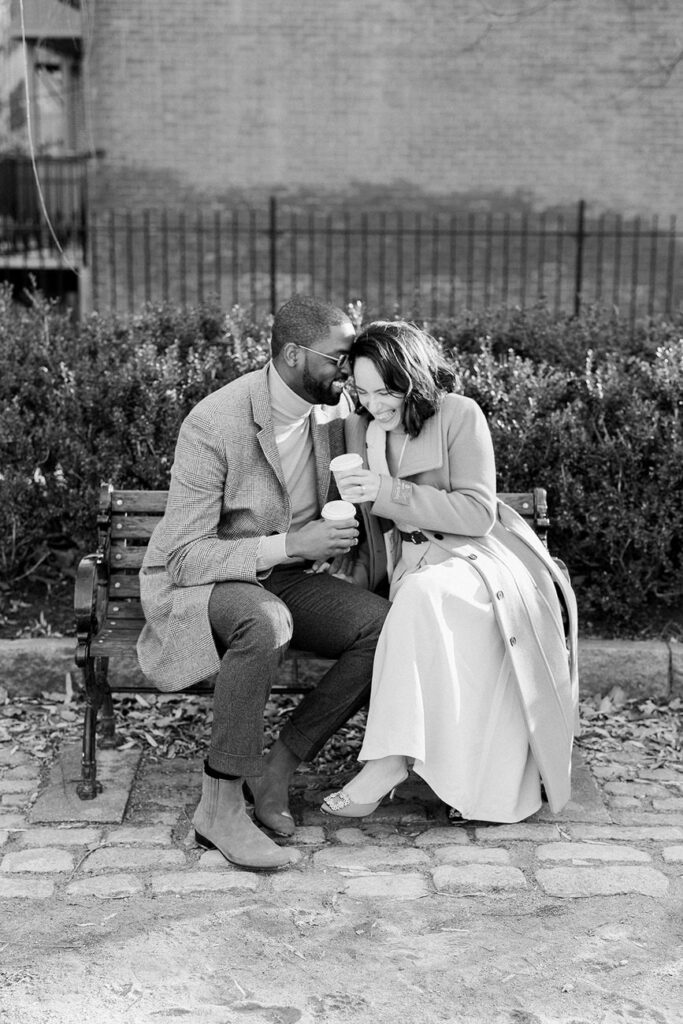 black and white photograph of a couple sitting close on a bench, sharing a laugh with a coffee cup in hand, with a textured wall and greenery in the backdrop.