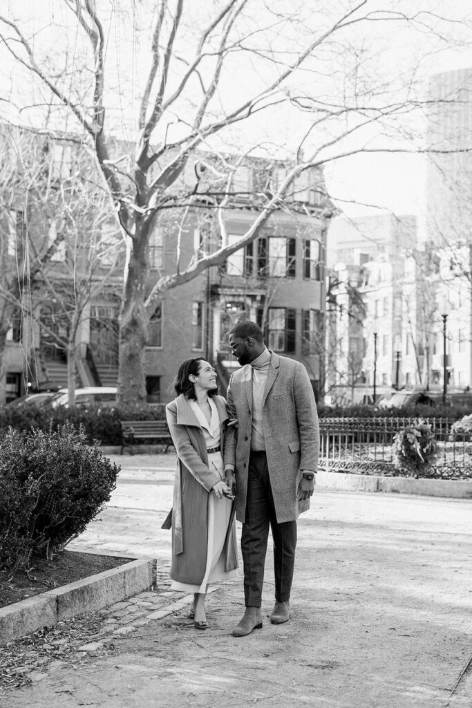 Black and white photo of a couple standing and holding hands in an urban park, with bare trees and historic buildings surrounding them.