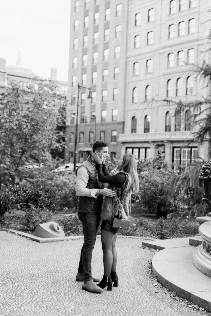 A couple embracing each other while standing on a cobblestone path in an urban park, with a backdrop of a classic apartment building