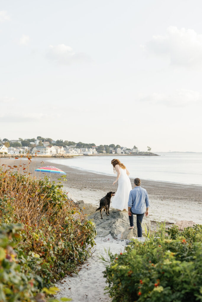 A couple climbing rocks on the beach, the man in a casual blue shirt and the woman in a white dress, with a happy dog looking back at them.