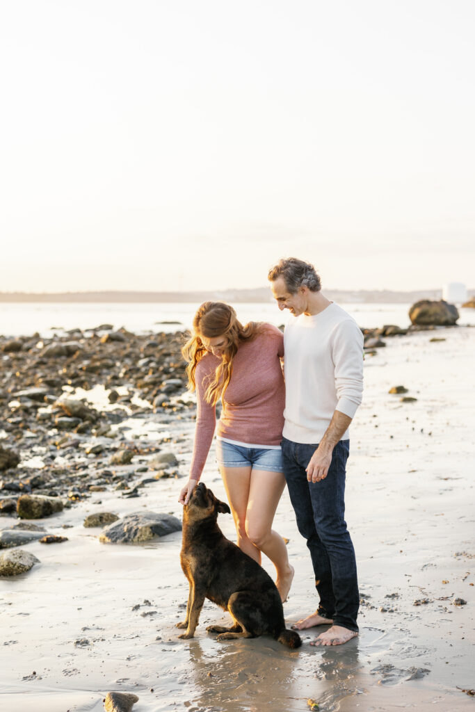 A couple with their dog at the beach, the woman in a pink sweater and denim shorts bending down to pet the dog, and the man in a white sweater looking on with affection.