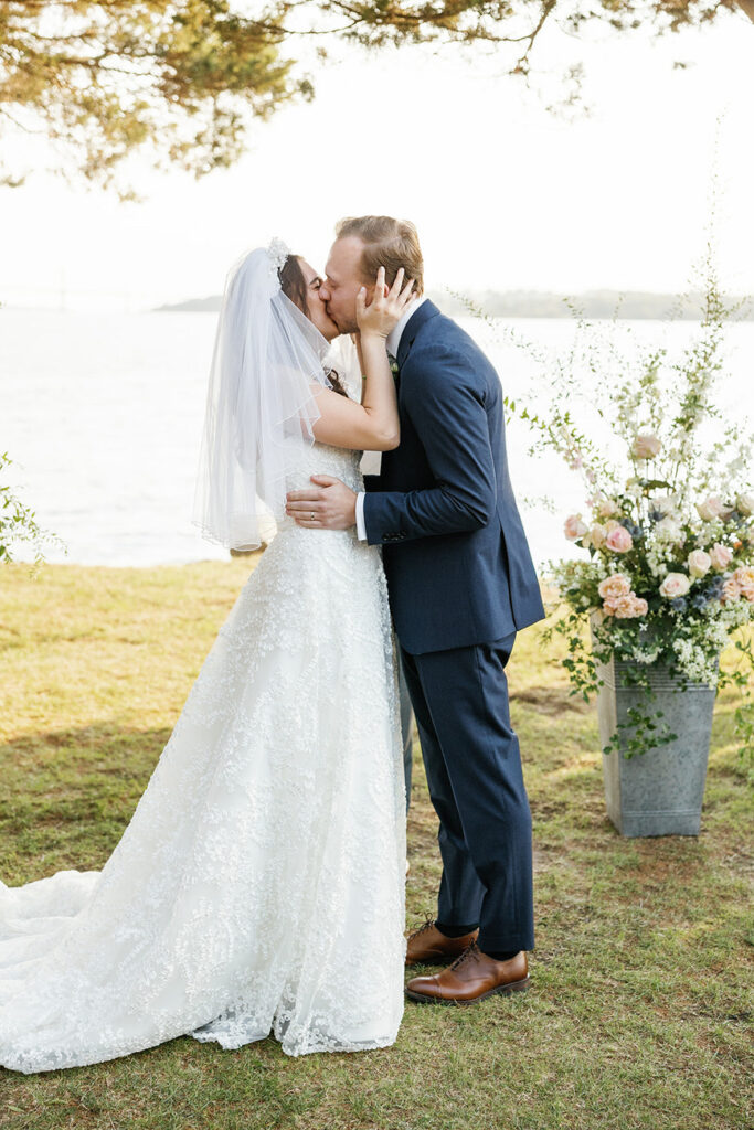 A bride and groom kiss passionately outdoors, with the bride's veil flowing and a floral arrangement in the background.