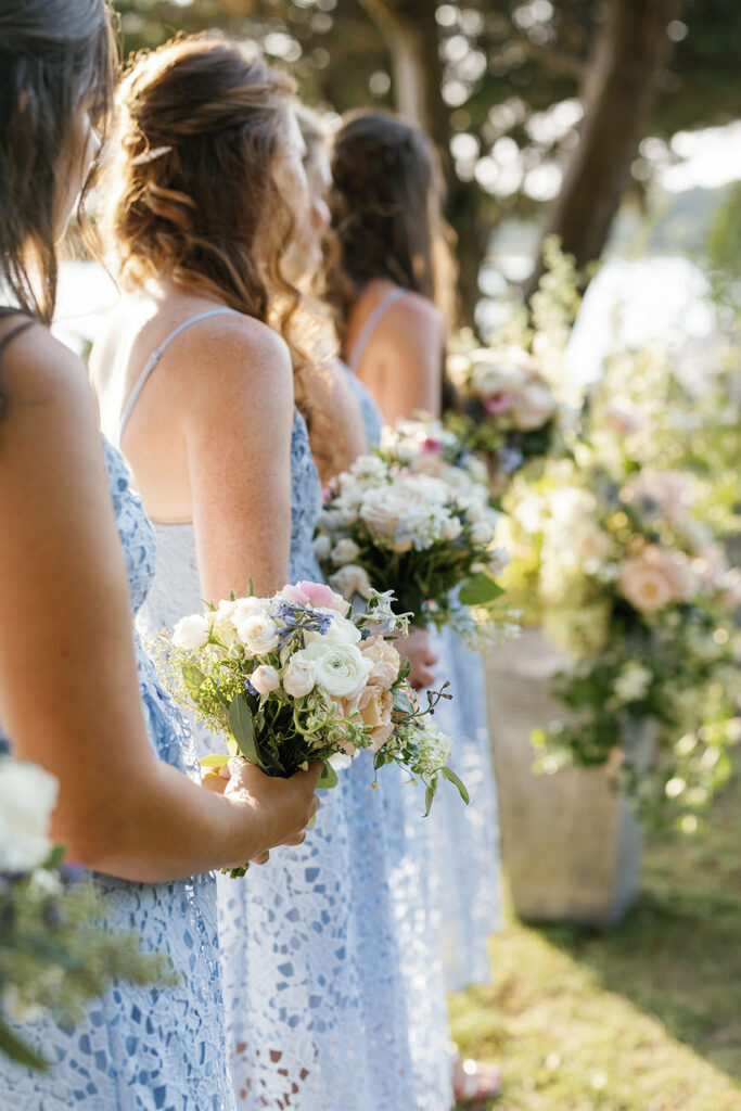 Bridesmaids in blue dresses hold bouquets of white and pink flowers, standing in a line during a wedding ceremony.