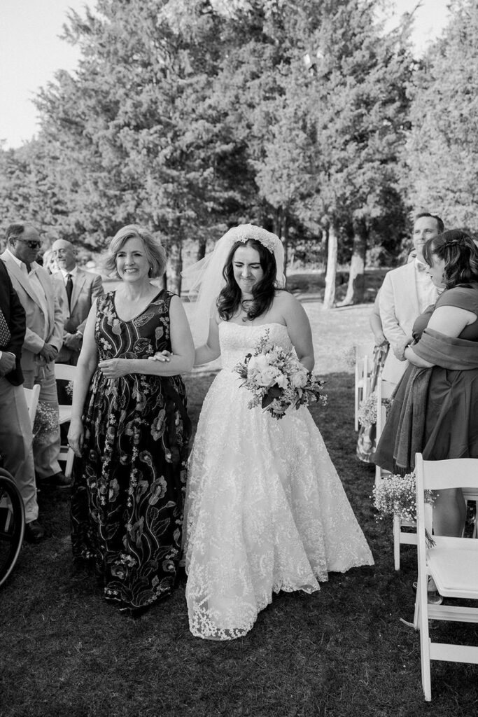 A bride walks down the aisle with an older woman, both smiling, in a black and white photograph.