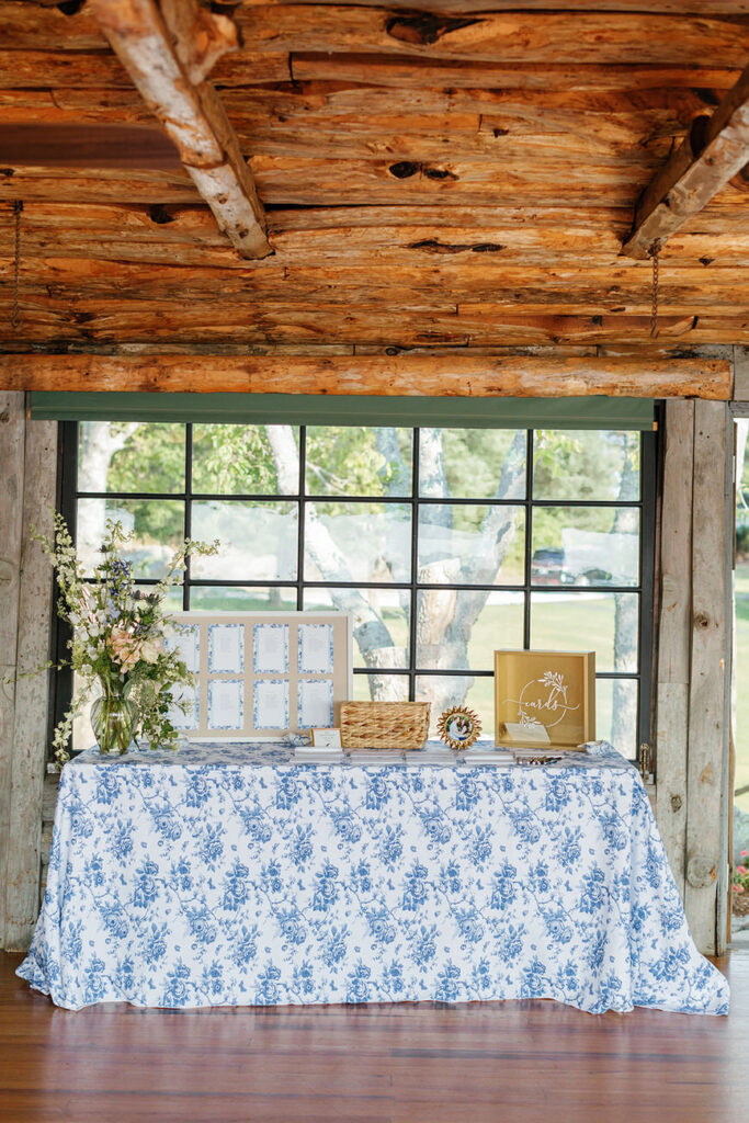 A rustic welcome table at a wedding, with a blue floral tablecloth and a variety of items including a seating chart, a basket, and a sign reading "cards," all under a wooden beam ceiling.