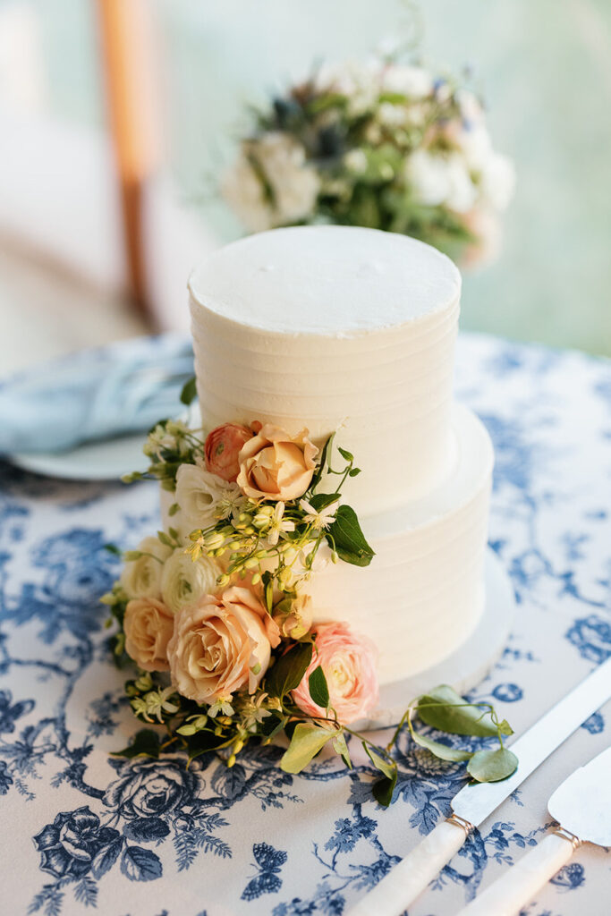 A simple white two-tiered wedding cake adorned with peach and cream roses, standing on a table with a blue floral tablecloth.