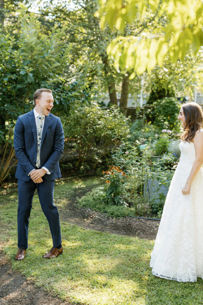 A groom in a blue suit laughs joyfully at his bride, in a lush green garden setting.