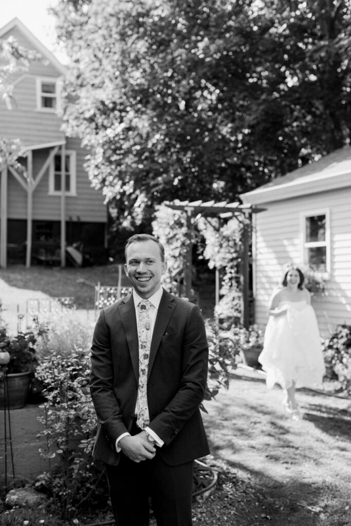A black and white photo capturing a smiling groom in the foreground, with the bride approaching from the background in a garden.