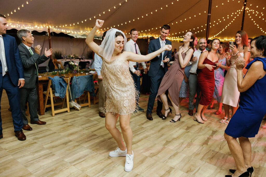 The bride, in a sparkling golden dress and sneakers, dances exuberantly among wedding guests under a canopy of string lights.