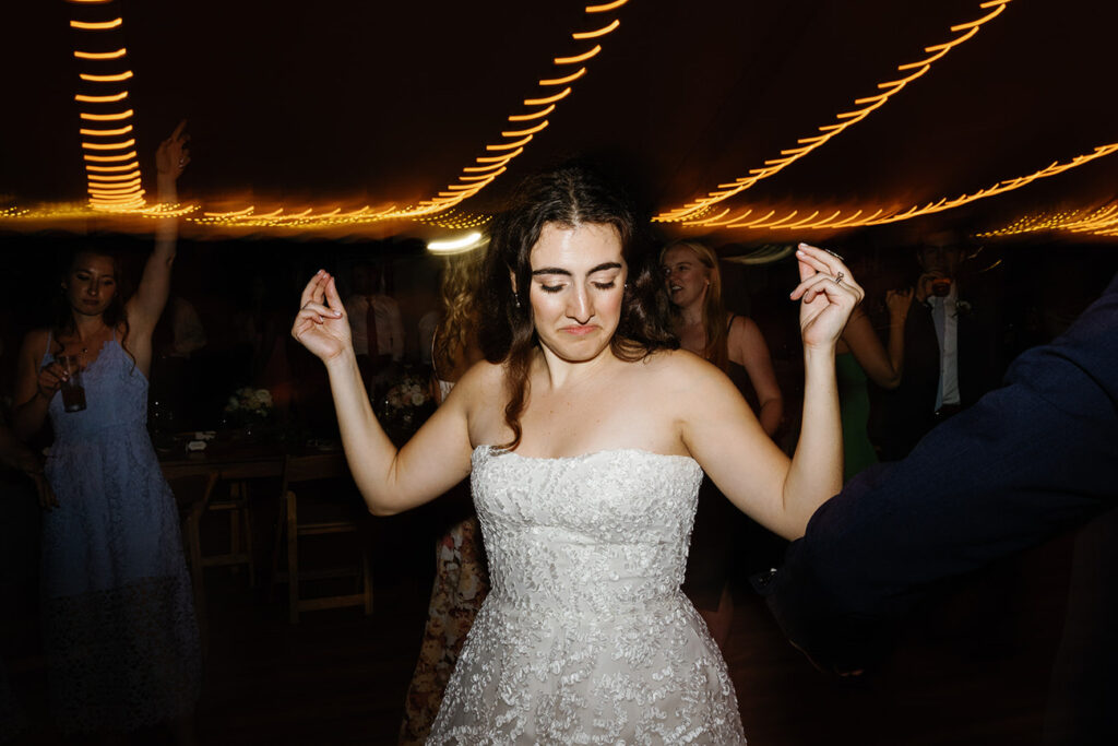 The bride, in a white wedding gown, dances with closed eyes, lost in the moment, during the reception under a tent with warm lighting.